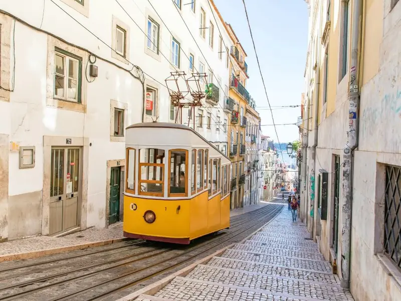 A classic yellow Lisbon tram travels along a steep cobbled street lined with historic buildings in Lisbon, Portugal, with a bright sky and a glimpse of the river in the distance. An iconic Lisbon city-break scene that fits an article on the safest places to visit in Europe, highlighting Portugal as a relaxed, visitor-friendly destination known for walkable neighbourhoods, easy public transport, and a generally low-stress atmosphere for mature travellers.