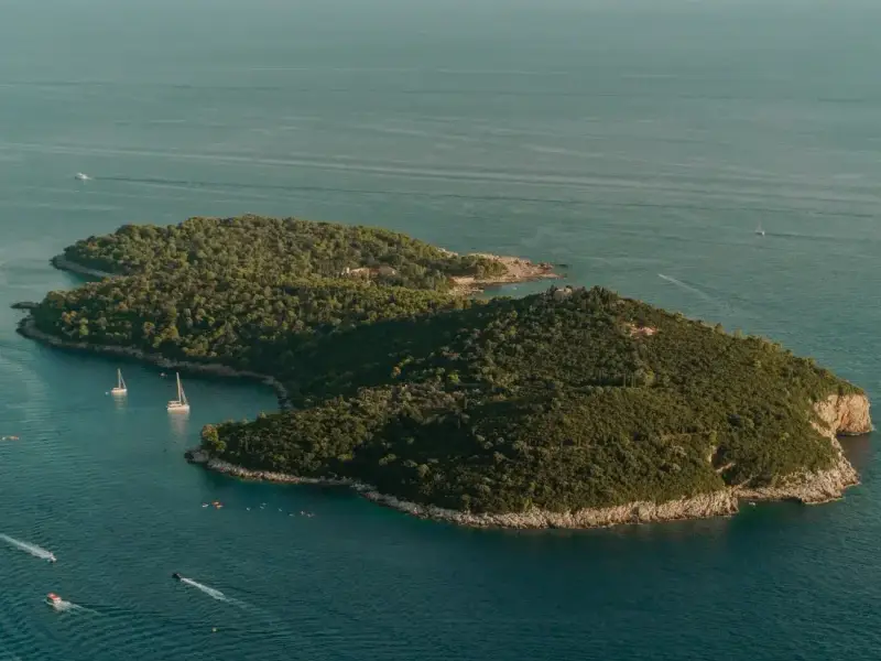 Aerial view of Lokrum Island just off the coast of Dubrovnik, Croatia, surrounded by deep blue Adriatic waters with sailboats nearby. Lokrum is one of the most popular day trips from Dubrovnik Old Town and offers forested walking paths, swimming spots and peaceful escapes from the busy city.