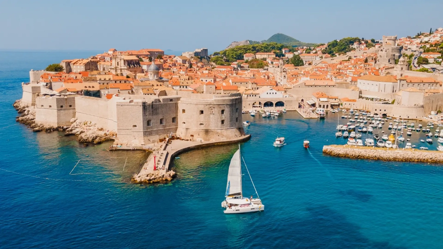 Aerial view of Dubrovnik Old Town on Croatia’s Adriatic coast, with stone city walls, terracotta rooftops and a small marina, while a sailing yacht crosses bright blue water — ideal for a 2026 Croatia islands and hotspots travel guide.