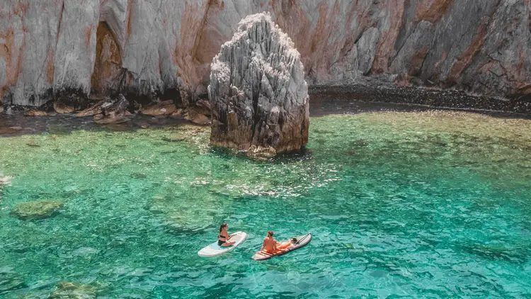 Two Yacht Getaways guests paddleboarding through crystal-clear turquoise waters in a secluded Mediterranean cove, surrounded by dramatic limestone cliffs and rocky coastal scenery.