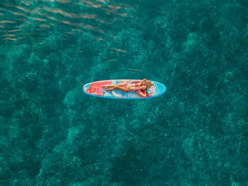 An overhead view of a traveller lying on a paddleboard in clear blue-green water, showing the calm and restorative side of a Mediterranean yacht holiday. The image reflects blue health travel through gentle movement, sunlight, open water and quiet moments at sea, ideal for a wellness-inspired catamaran cruise in Greece or Croatia.