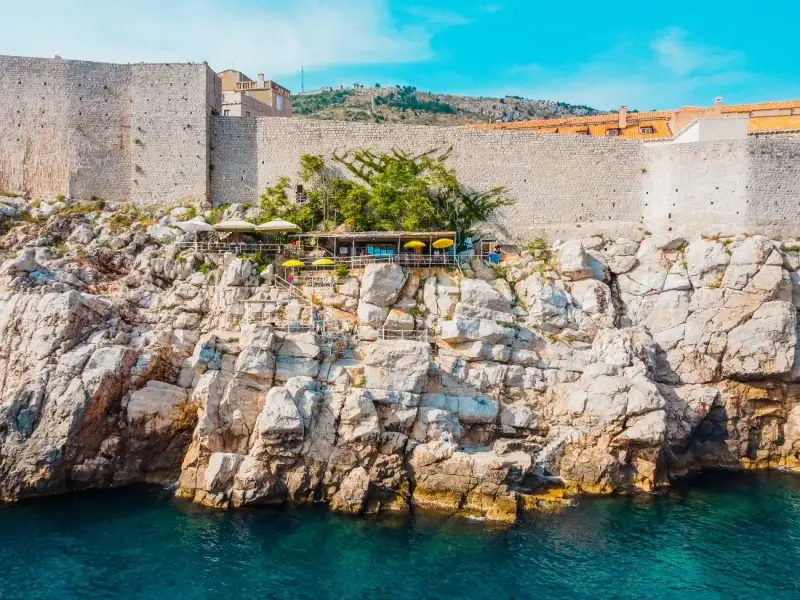 Stone city walls of Dubrovnik rising above the rocky Adriatic coastline in Croatia with a seaside terrace and clear blue water below. Dubrovnik’s famous fortifications are among the best preserved medieval walls in Europe and are one of the most iconic sights in the historic Old Town.
