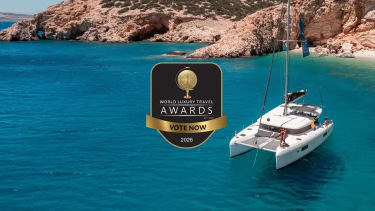 Luxury Yacht Getaways catamaran anchored in a secluded Mediterranean bay beside rocky Greek islands and turquoise water, featuring the 2026 World Luxury Travel Awards Vote Now badge following Yacht Getaways’ nomination for Best Yacht Cruises.