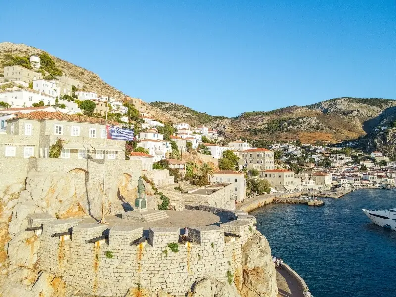 Hydra harbour in Greece with stone cannon terrace, hillside houses, and boats on calm blue water.