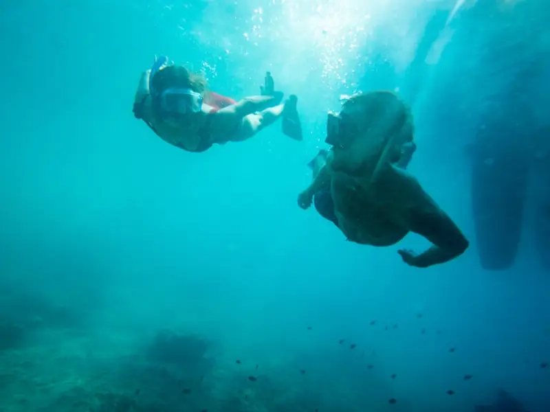 Underwater photo of two swimmers snorkelling beneath the surface near Glyka Nera, also known as Sweetwater Beach, on the south coast of Crete. Known for its clear blue water and dramatic remote setting, this beautiful beach is one of the most memorable boat-only stops for a swimming or snorkelling break during a sailing trip in Greece.