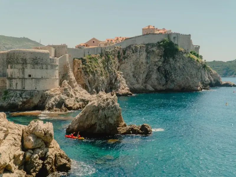 Historic Dubrovnik city walls rising above the rocky Adriatic coastline in Croatia, with turquoise water below and kayakers exploring the shoreline near the UNESCO-listed Old Town. This coastal perspective highlights the dramatic setting of Dubrovnik and why exploring the city walls and surrounding sea is one of the most memorable experiences in a Dubrovnik travel guide.