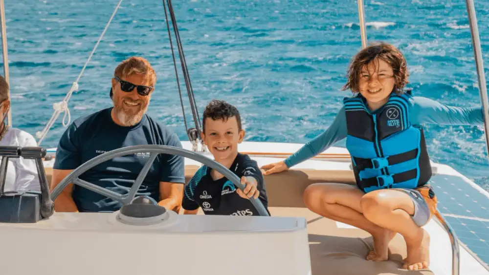 Two smiling children enjoying a hands-on sailing experience aboard a Yacht Getaways catamaran, with one steering the yacht alongside the professional skipper while the other sits nearby wearing a life jacket.