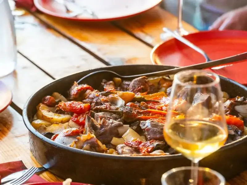 A rustic meal served on a wooden table in Dubrovnik, Croatia, with a glass of white wine and a generous dish of roasted vegetables and hearty local flavours in the traditional Peka dish. This image adds to a Dubrovnik travel guide by highlighting one of the best parts of visiting the city: enjoying long, relaxed meals, local food and the more refined culinary side of a Croatia holiday.