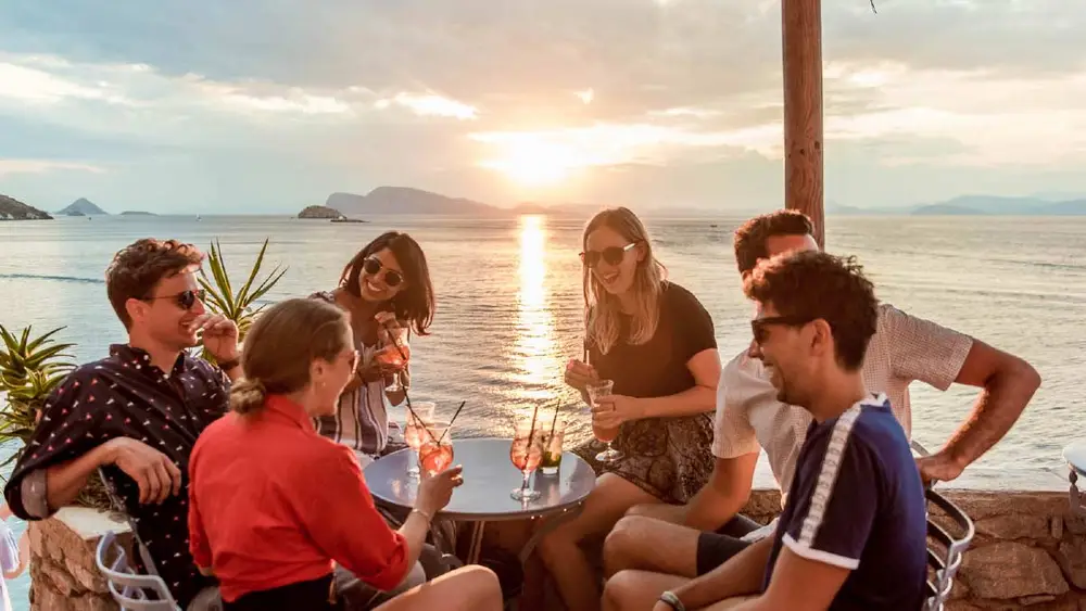 A group of people have sunset cocktails and laugh and talk while at a seaside bar in Lefkada Greece on a luxury sailing holiday.