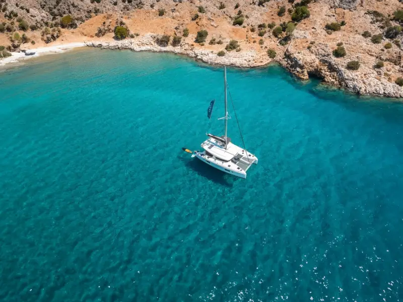 Aerial view of a catamaran anchored in a quiet bay off Kato Koufonisi in the Cyclades, surrounded by crystal-clear turquoise water and a rugged, unspoilt coastline. This secluded Greek bay is only accessible by boat, making it a dream stop for sailing holidays in Greece and travellers looking for hidden beaches away from the crowds.