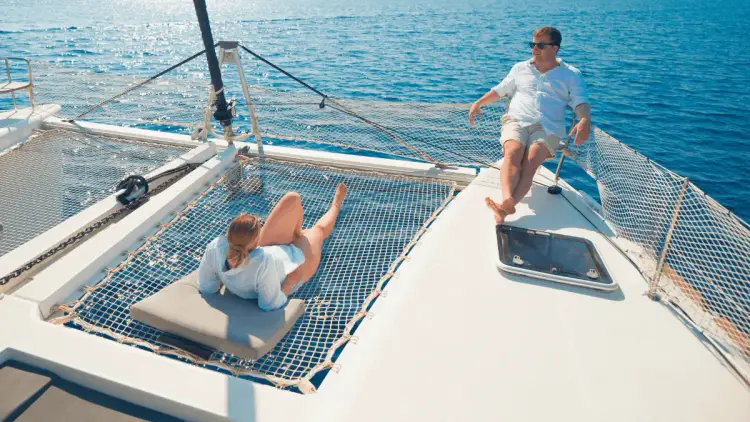 Couple relaxing in the Mediterranean sunshine on the spacious bow of a premium sailing catamaran, lounging on the deck and trampoline netting above the blue sea.