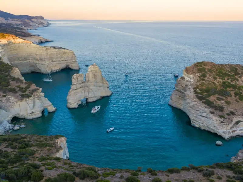 Aerial view of Kleftiko Bay on Milos, where white volcanic cliffs and rock formations rise above vivid blue water filled with anchored boats. Only accessible by sea, Kleftiko is one of the most famous bays in Greece for boat trips, swimming, and exploring sea caves, and it is a standout highlight of any Cyclades sailing holiday.