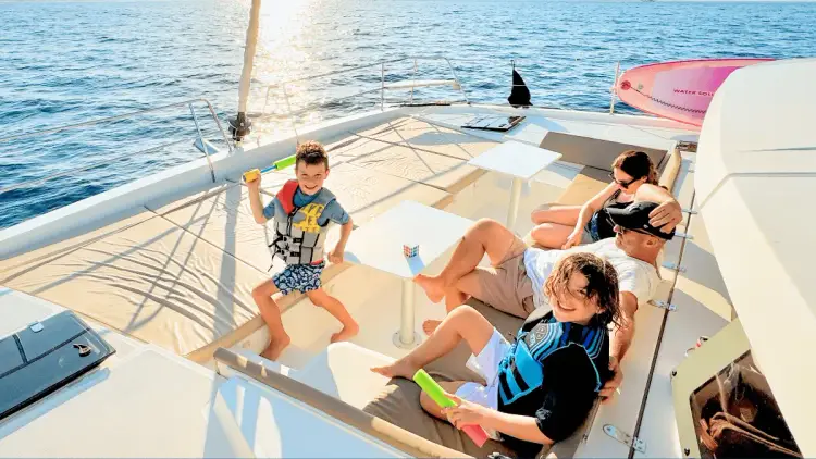 Family of four enjoying time together on the spacious outdoor lounge of a premium Yacht Getaways catamaran, with two children in life jackets playing while their parents relax in the Mediterranean sunshine.