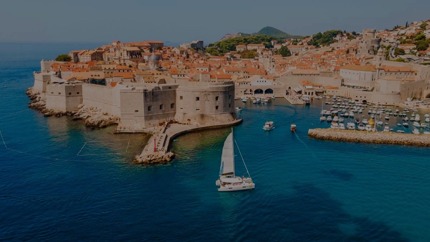 Aerial photo of Dubrovnik old town and fortifications with bright blue waters and a Yacht Getaways luxury catamaran sailing by.