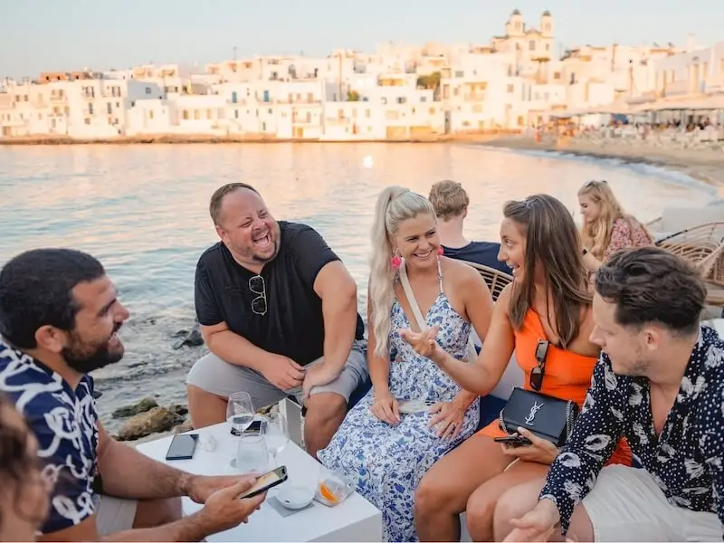 Photo of a group of people laughing and dining seaside on a picturesque Greek island in the Cyclades while on a luxury catamaran tour with Yacht Getaways.