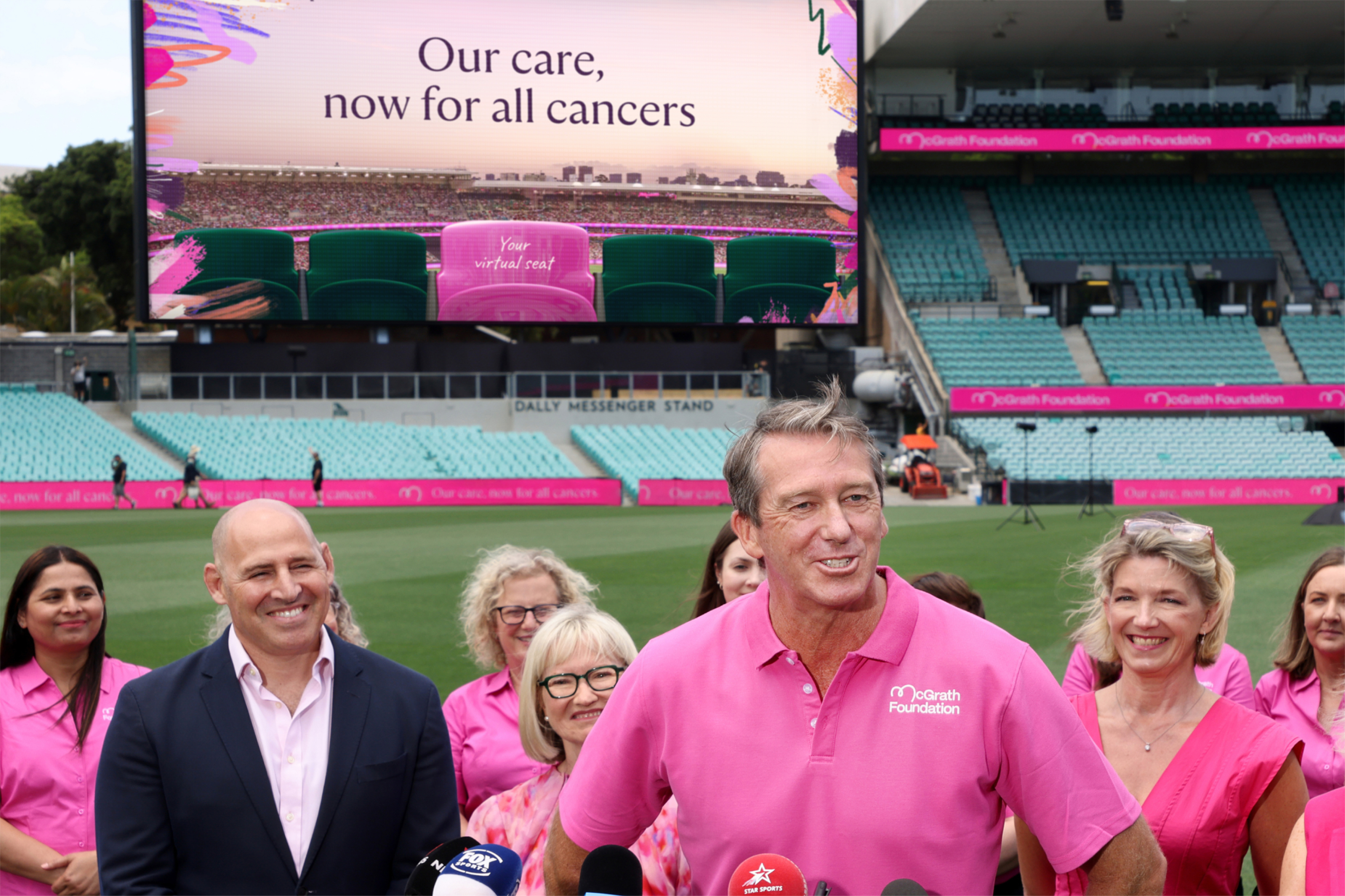 Launch event at the cricket ground, pink-shirted supporters under a screen reading Our care, now for all cancers