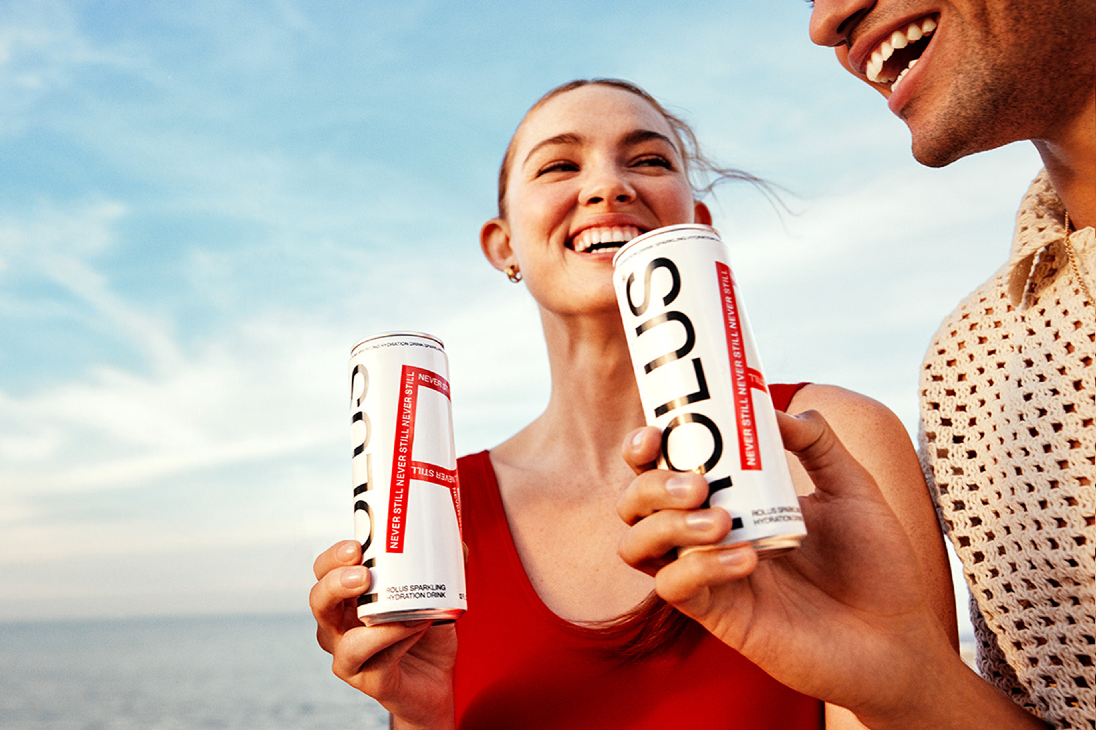 Two people laughing with cans by the sea, red swimsuit against a pale blue sky