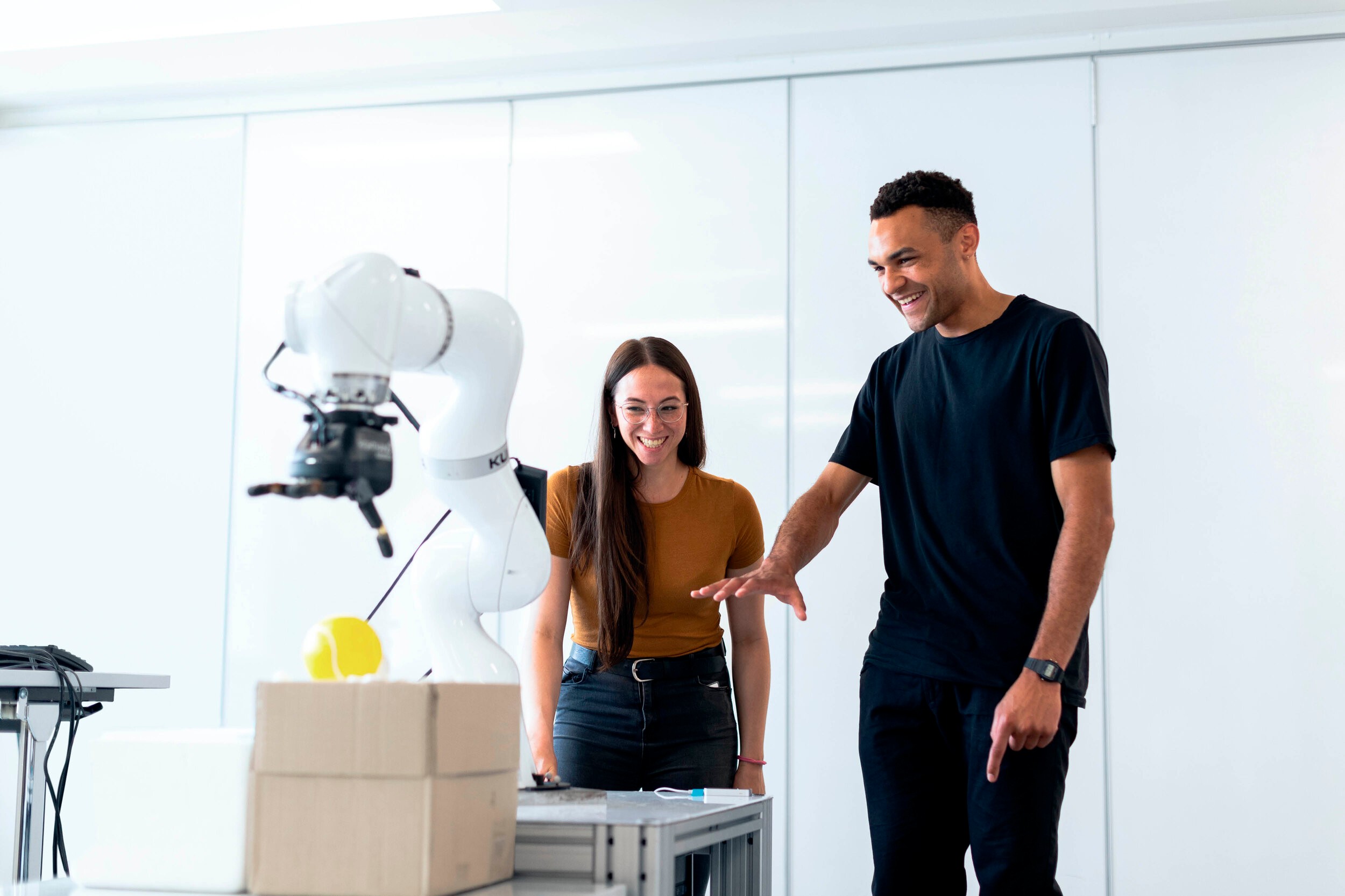 Two researchers laughing beside a white robotic arm in a bright, minimal lab space