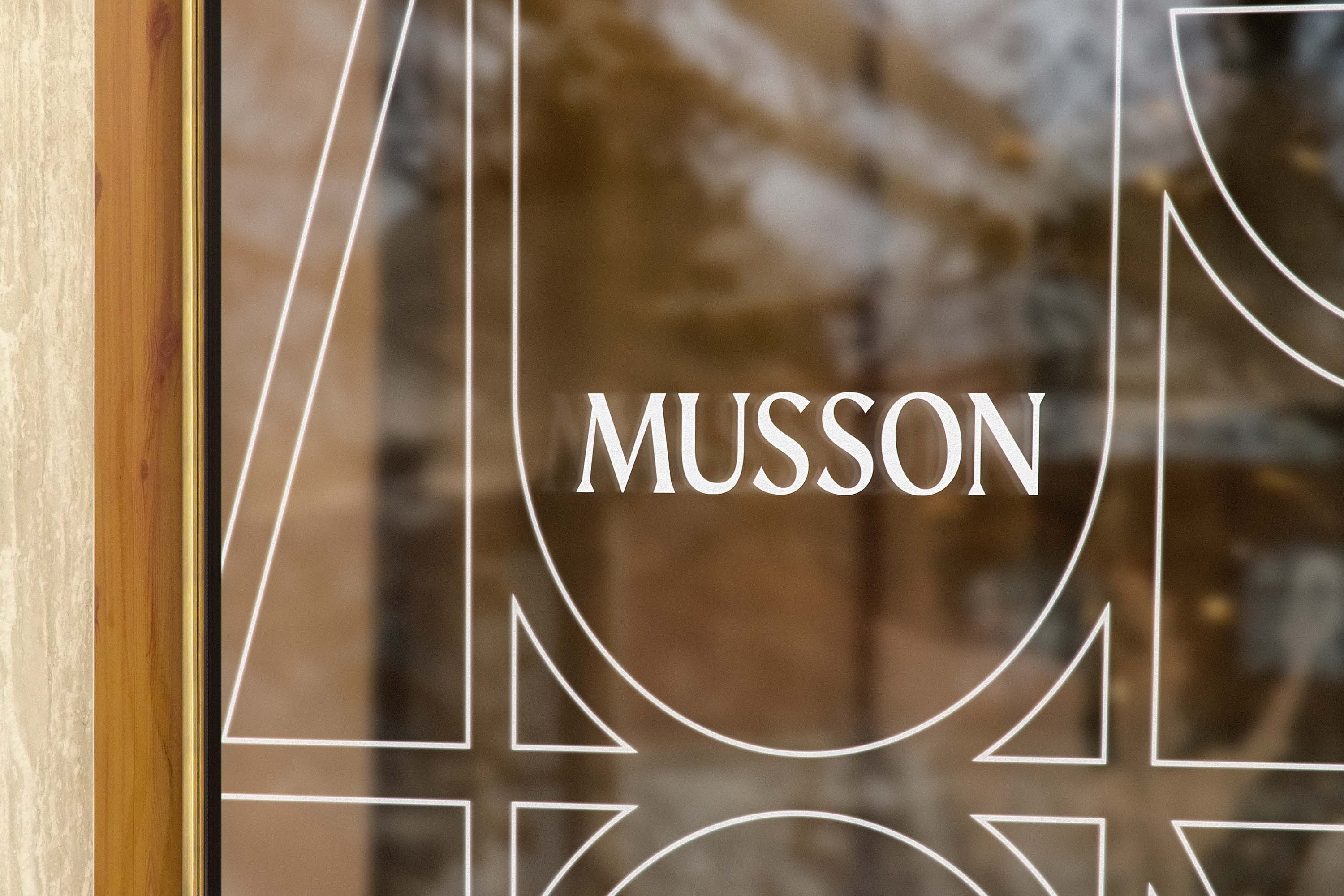 Etched glass shopfront window carrying white outline monogram lines and the Musson wordmark