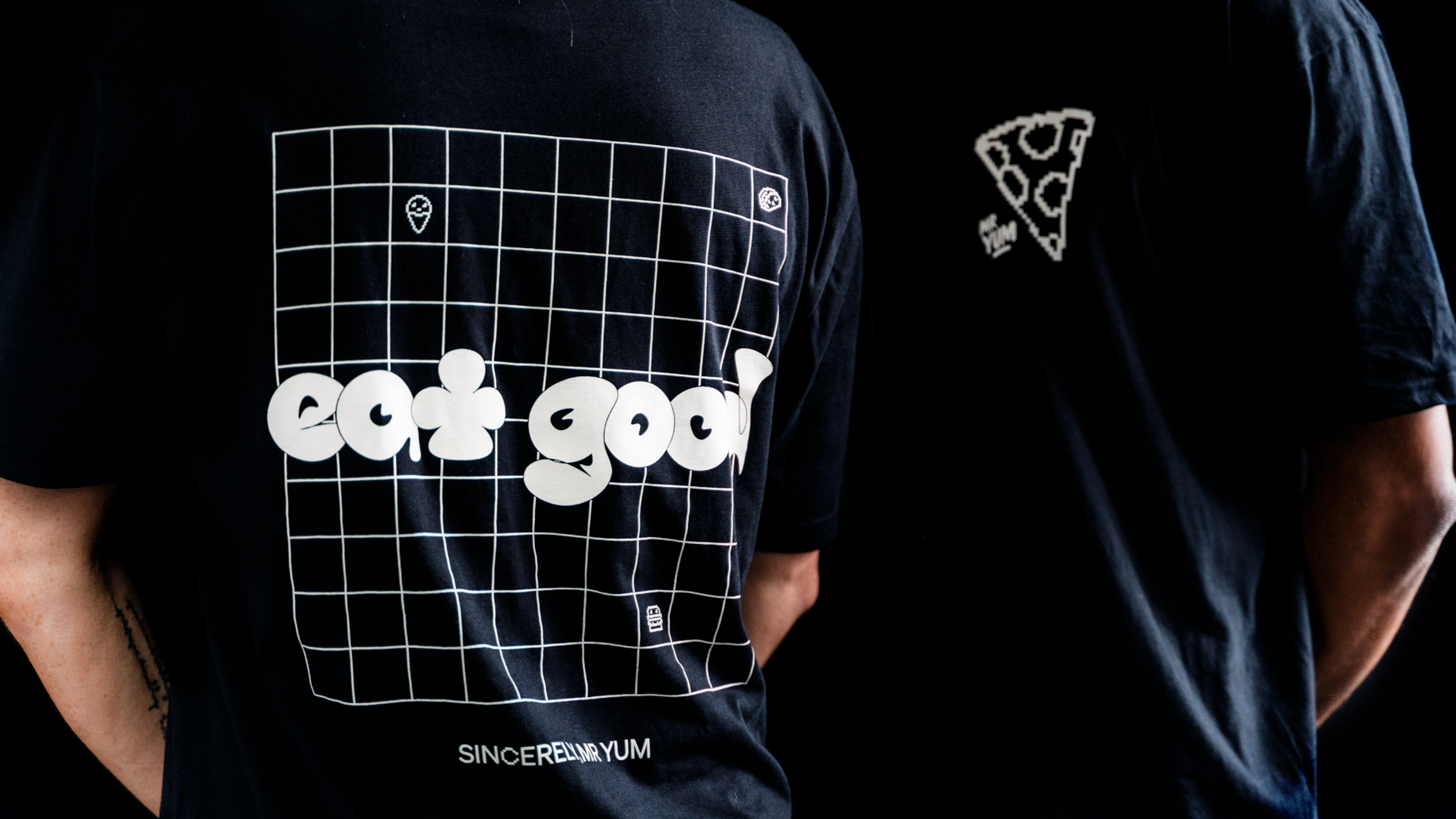 Backs of two black tees with bubble 'eat good' lettering on a wireframe grid and a pizza motif