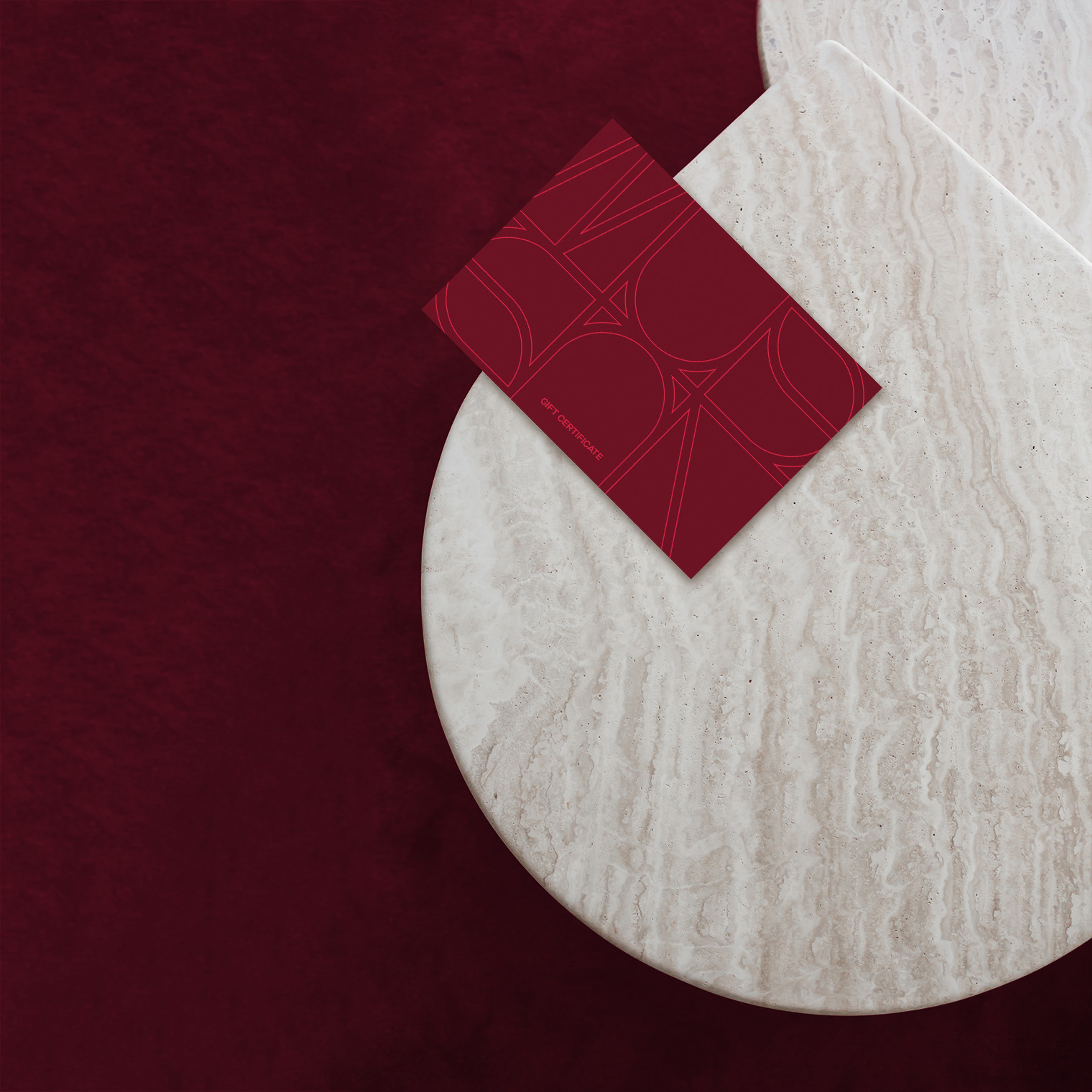 Burgundy gift card with an outlined monogram pattern on a travertine table over red velvet