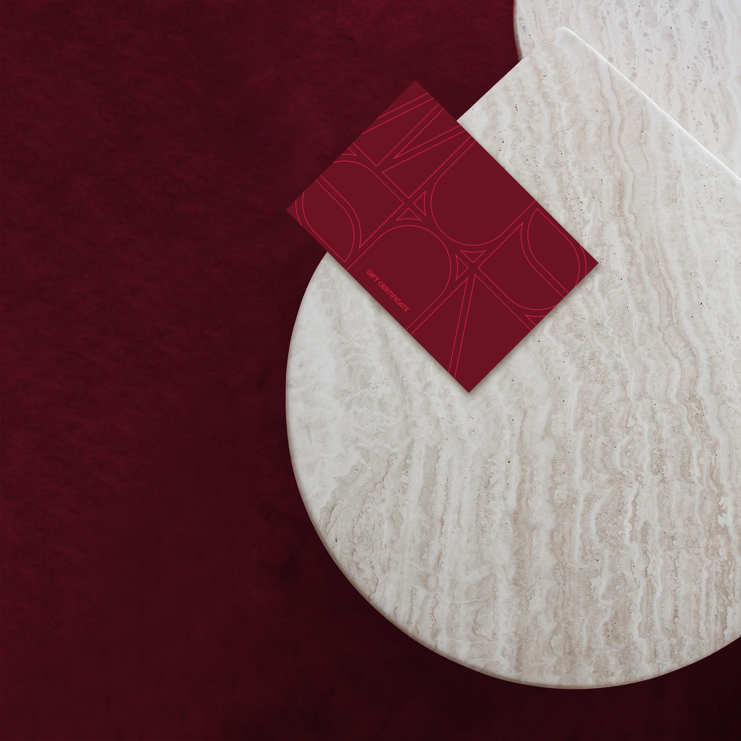 Burgundy gift card with an outlined monogram pattern on a travertine table over red velvet