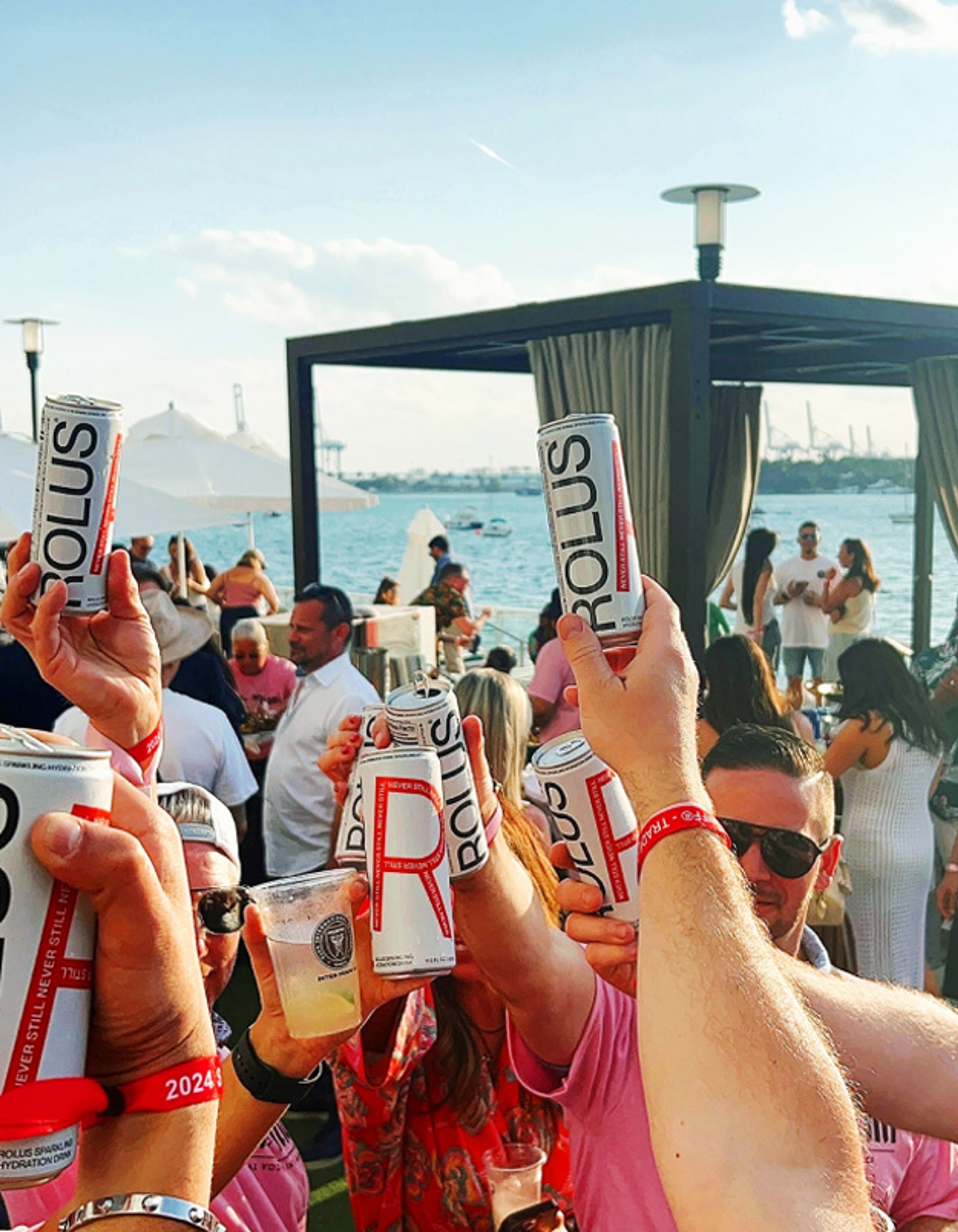Cans raised in a toast above a crowd at a waterfront event, sea and cabana behind