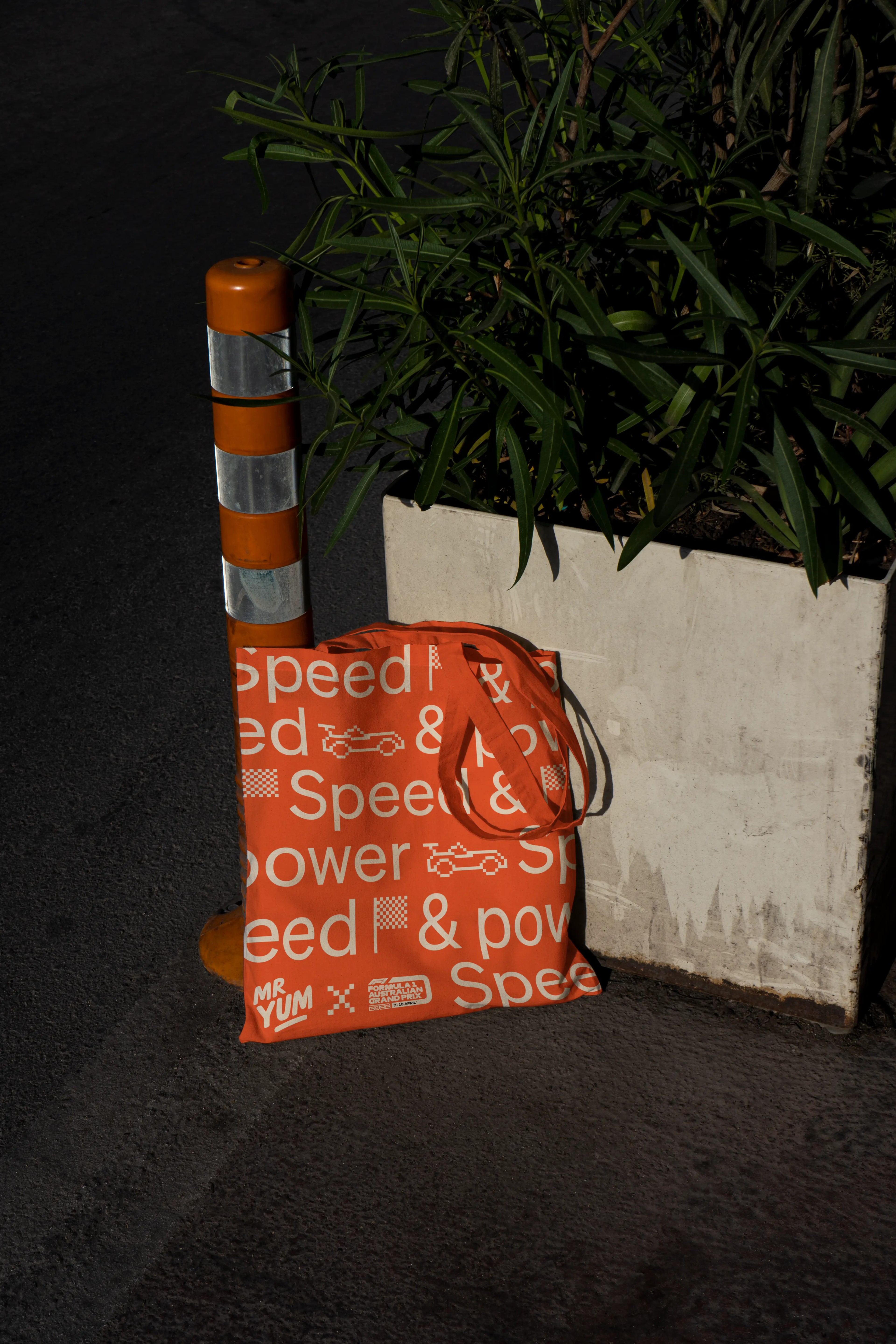 Orange Mr Yum tote printed with repeating 'Speed & power' type, propped against a road cone