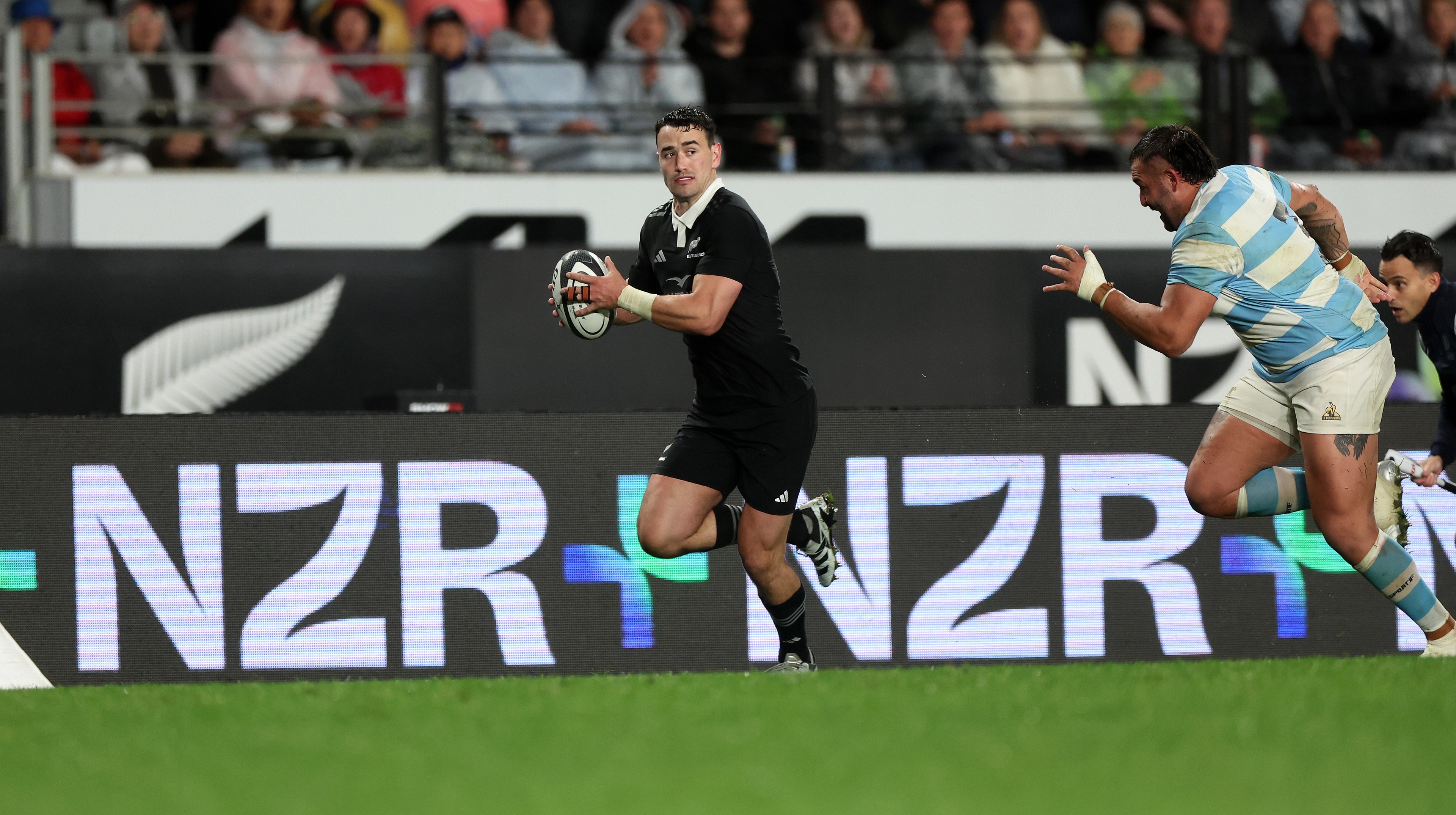 All Blacks player breaking clear of a defender in blue and white, NZR+ LED boards ringing the pitch