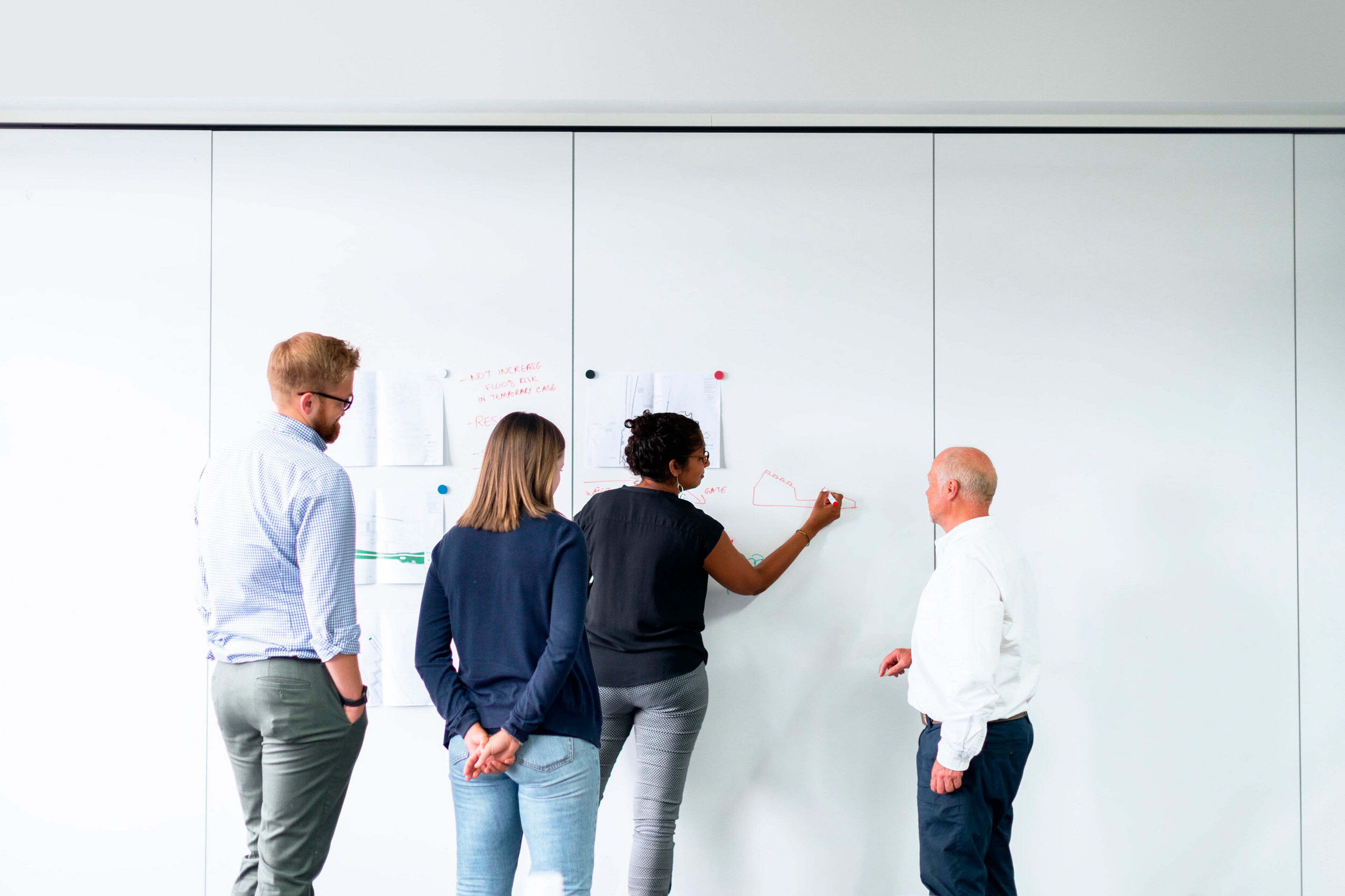 Four colleagues studying pinned notes and sketches on a floor-to-ceiling whiteboard wall