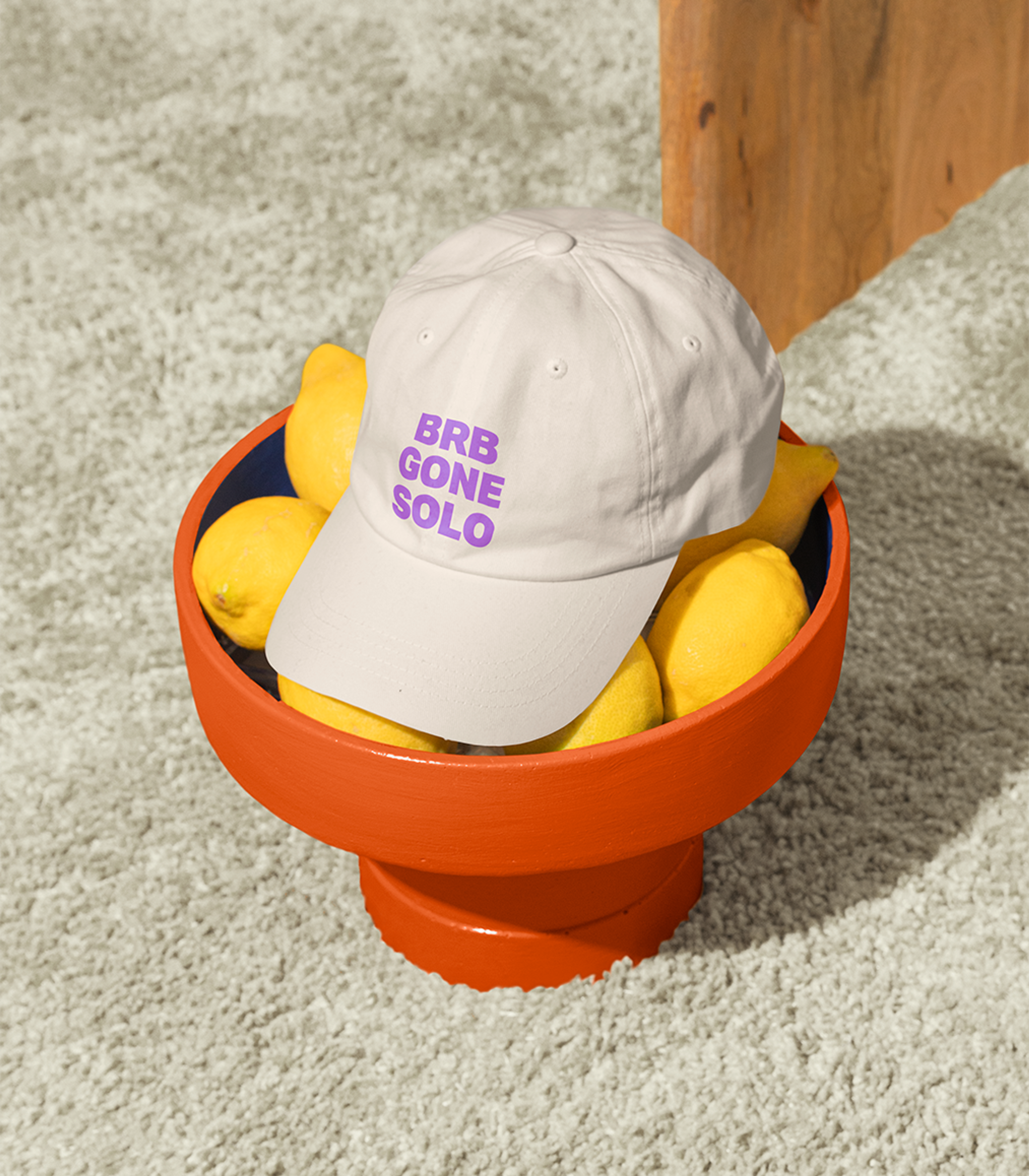 Cream cap embroidered 'BRB gone solo' in purple, perched on an orange bowl of lemons