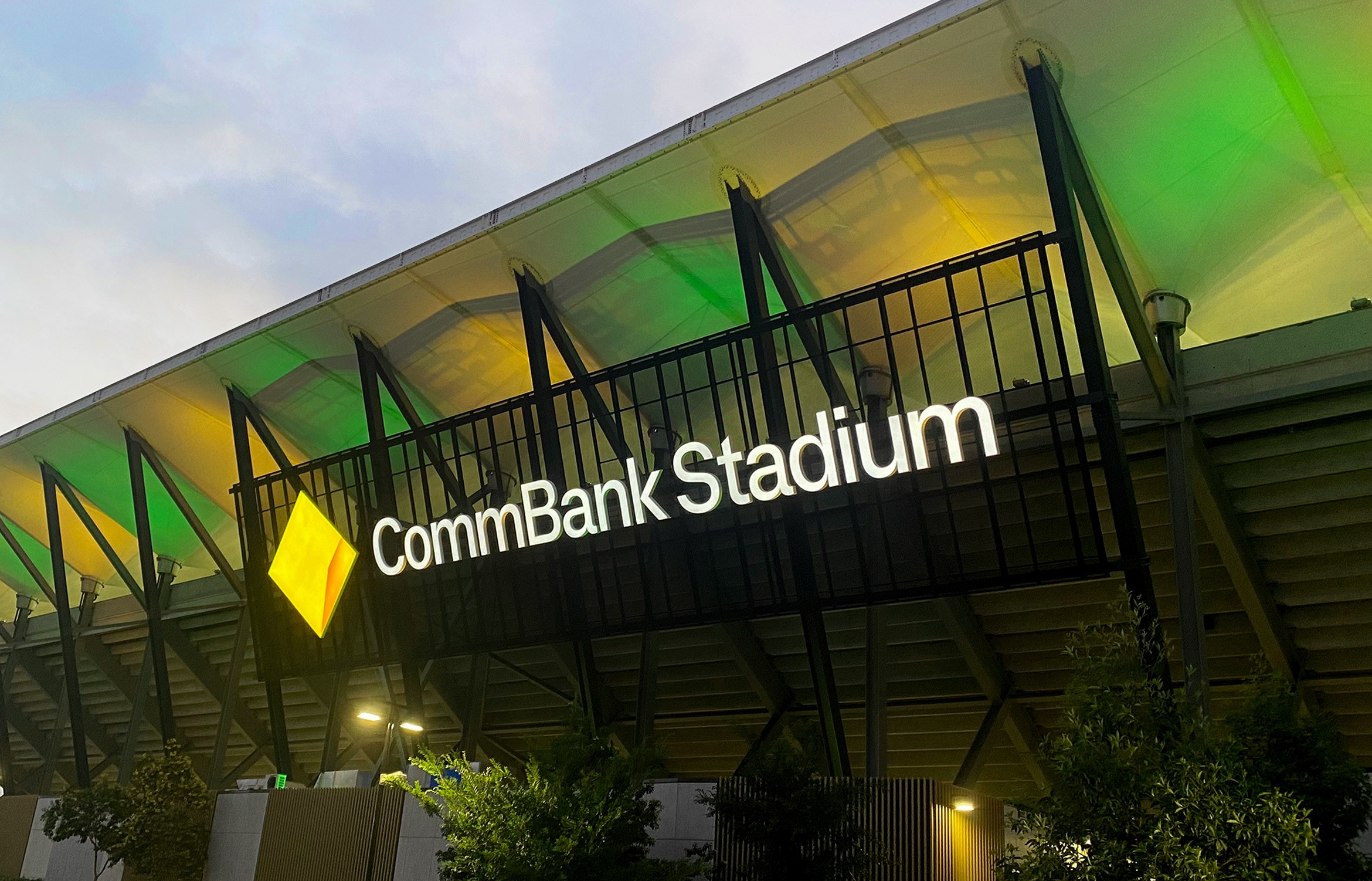 Illuminated CommBank Stadium signage and yellow diamond, roof lit green at dusk