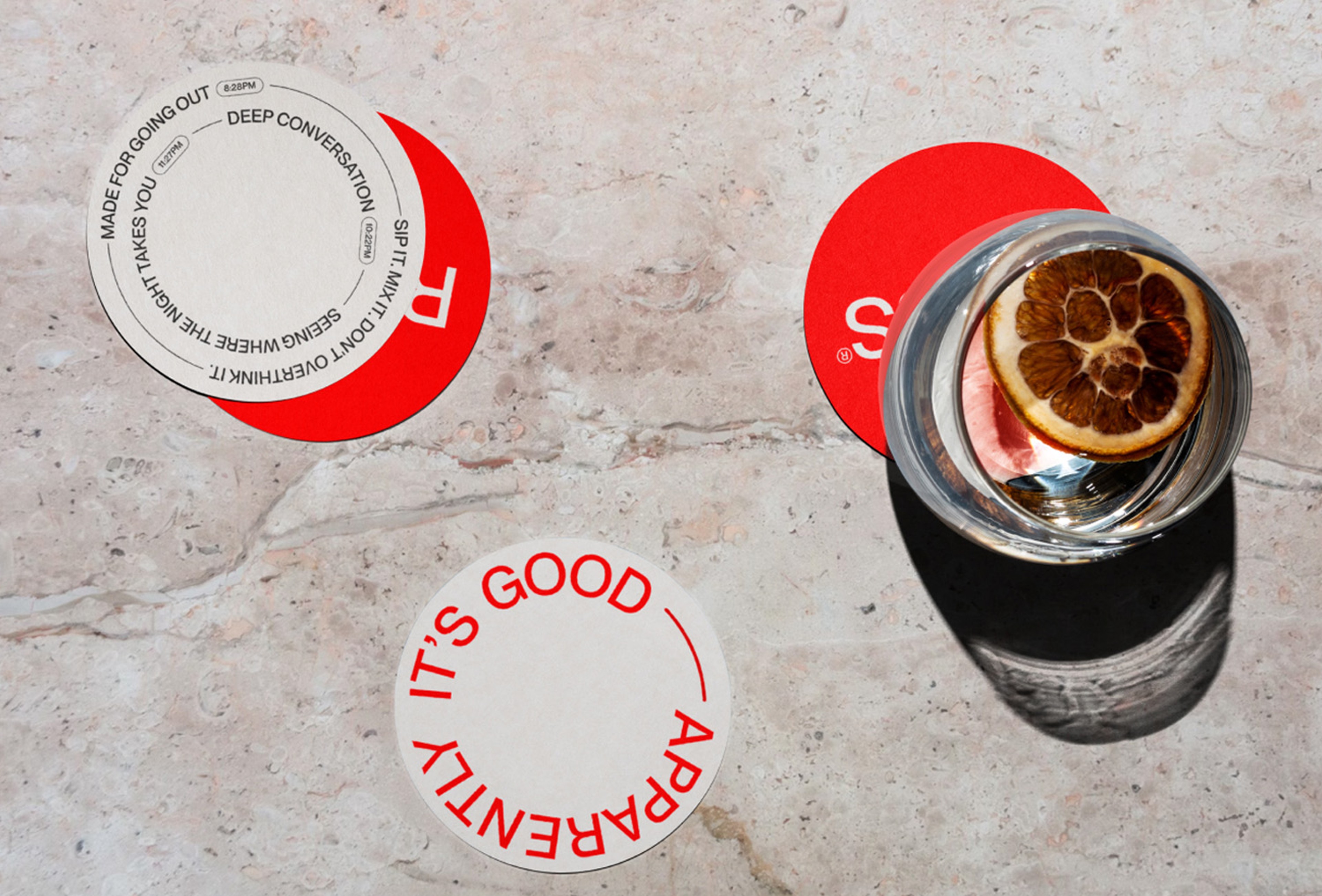 Rolus coasters in red and off-white with slogans set in circles, beside a citrus-topped drink