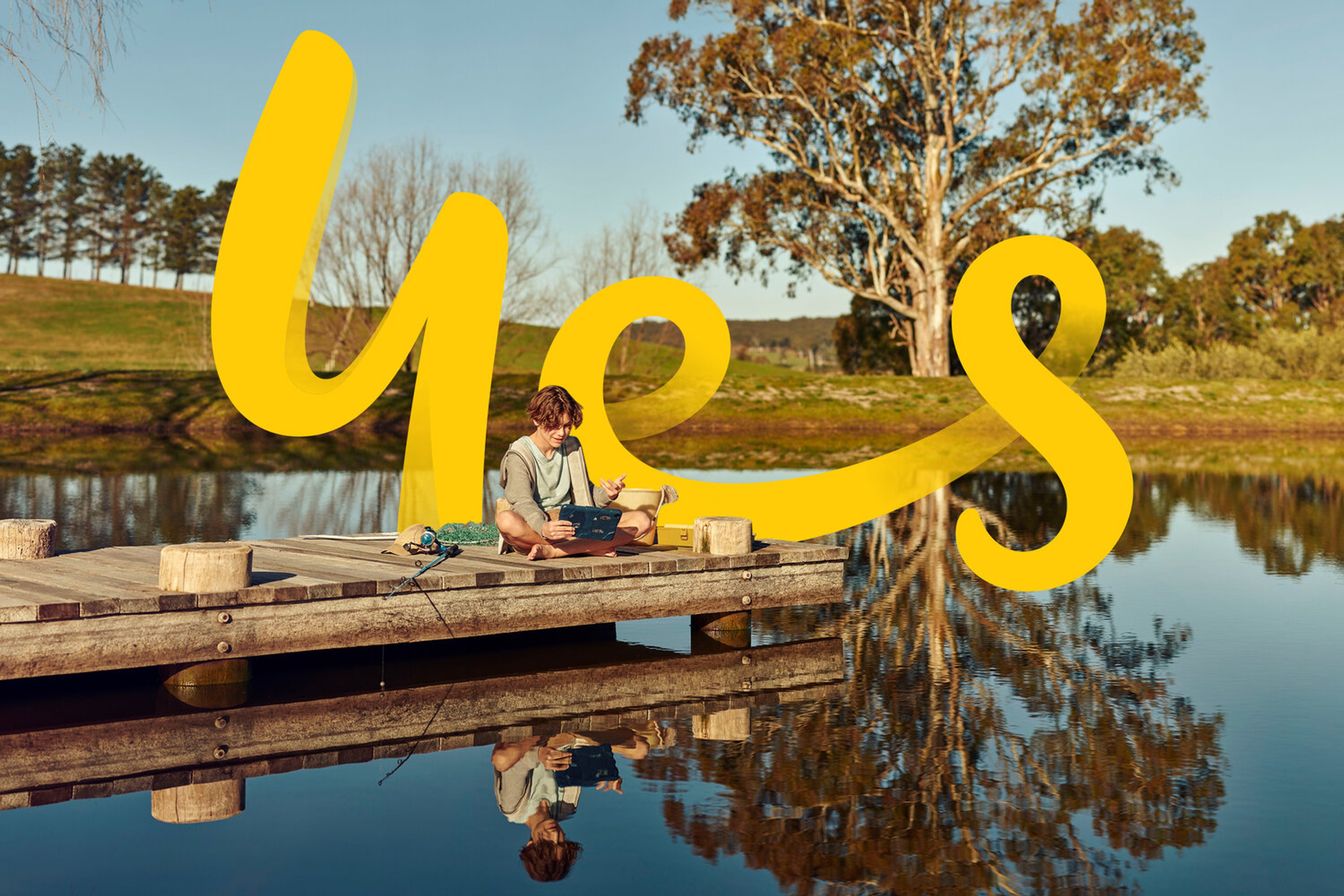 Lakeside jetty scene with the yellow 'yes' signature drawn large across the landscape