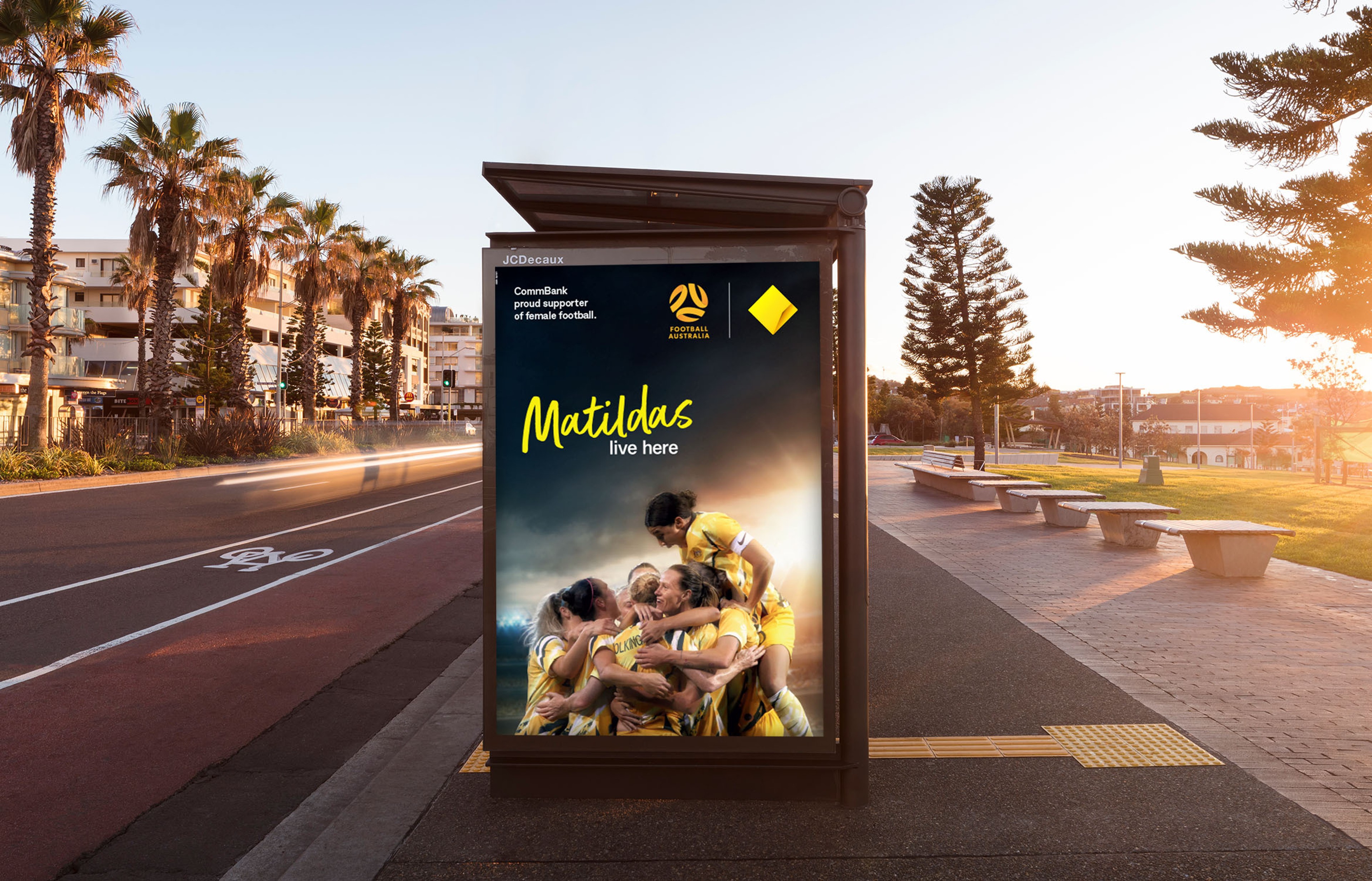 Seaside bus shelter panel carrying the CommBank Matildas football poster at sunset
