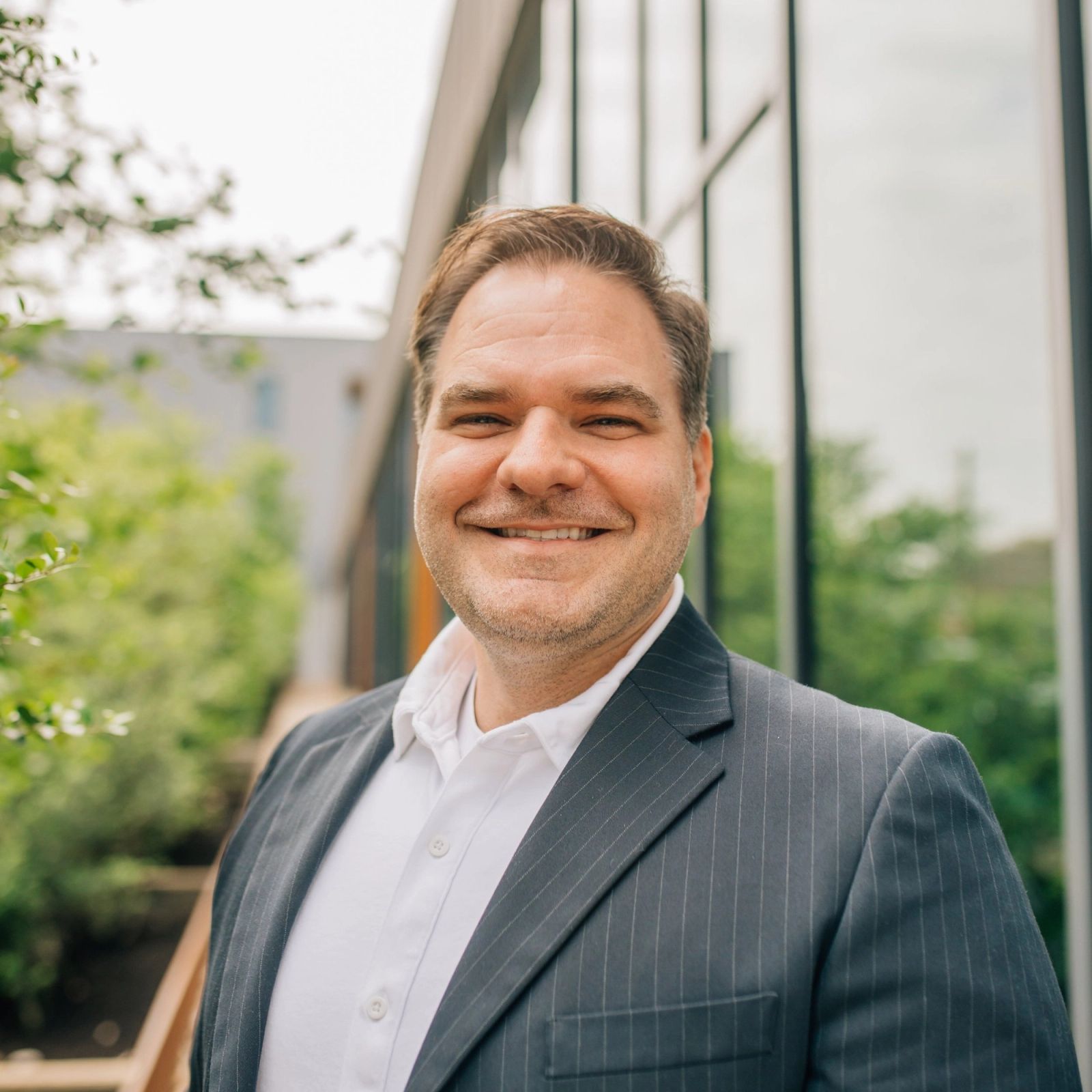 Chris in a suit and white shirt is smiling in front of a building .