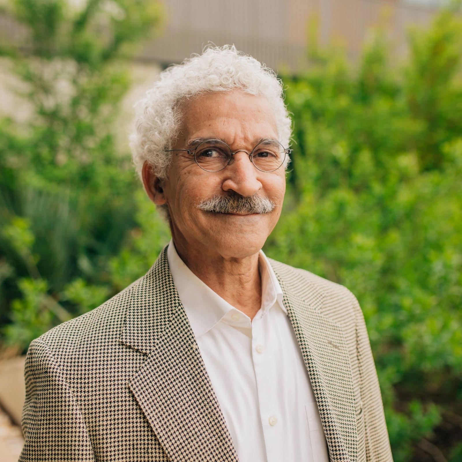 a man with glasses and a mustache is standing in front of a green bush .
