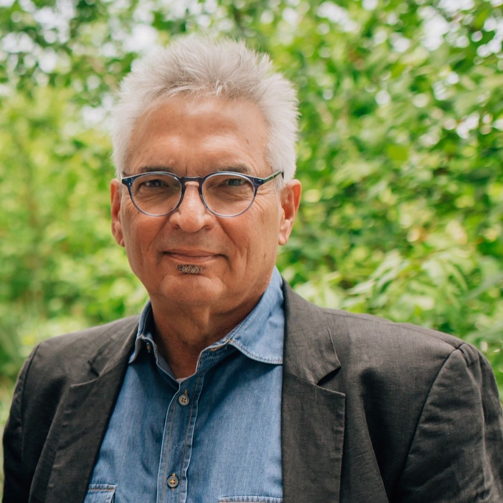 a man wearing glasses and a jacket is standing in front of a tree .