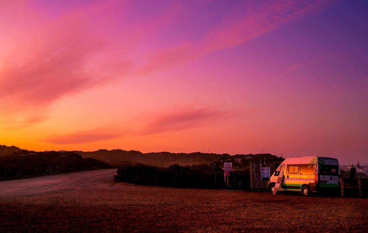 Camper van with a surfboard parked at a coastal campsite under a pink and orange sunset in Stillbaai