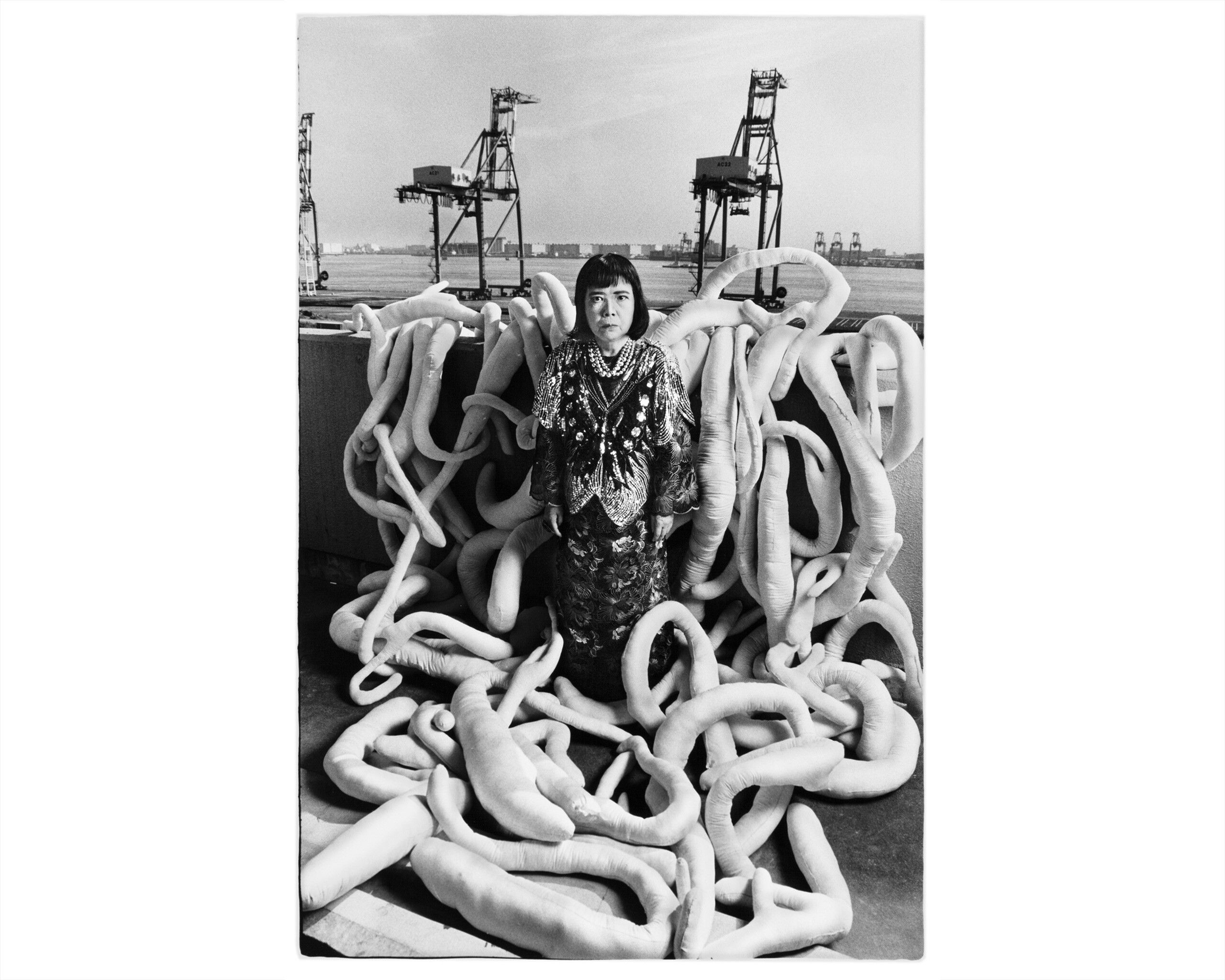 A black and white photo with Yayoi Kusama sitting in the middle of her sculpture work near a pier, dated 1993