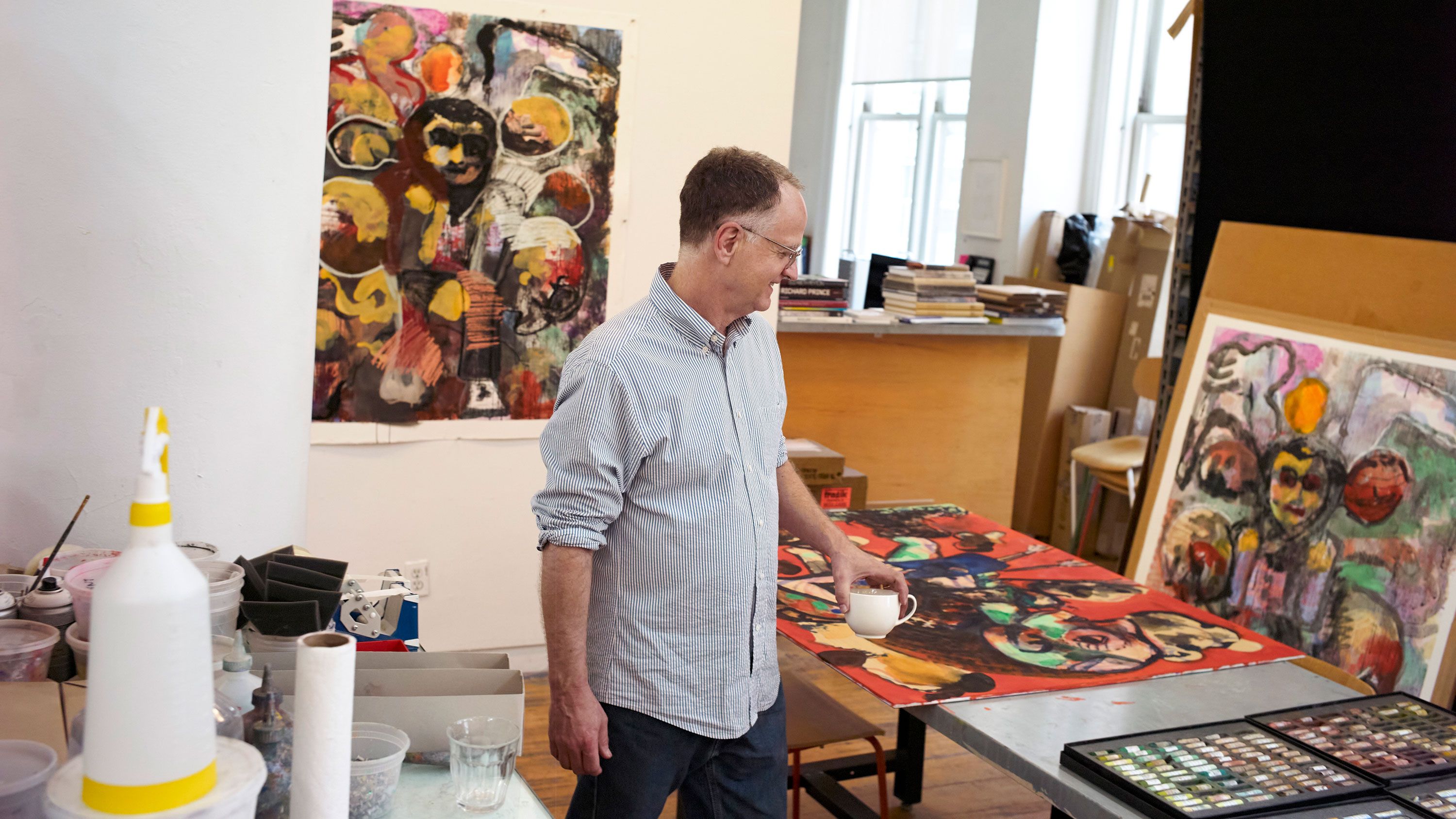 Peter Doig in his studio with the background of his prints hanging on the wall and laying on the desk, dated 2021