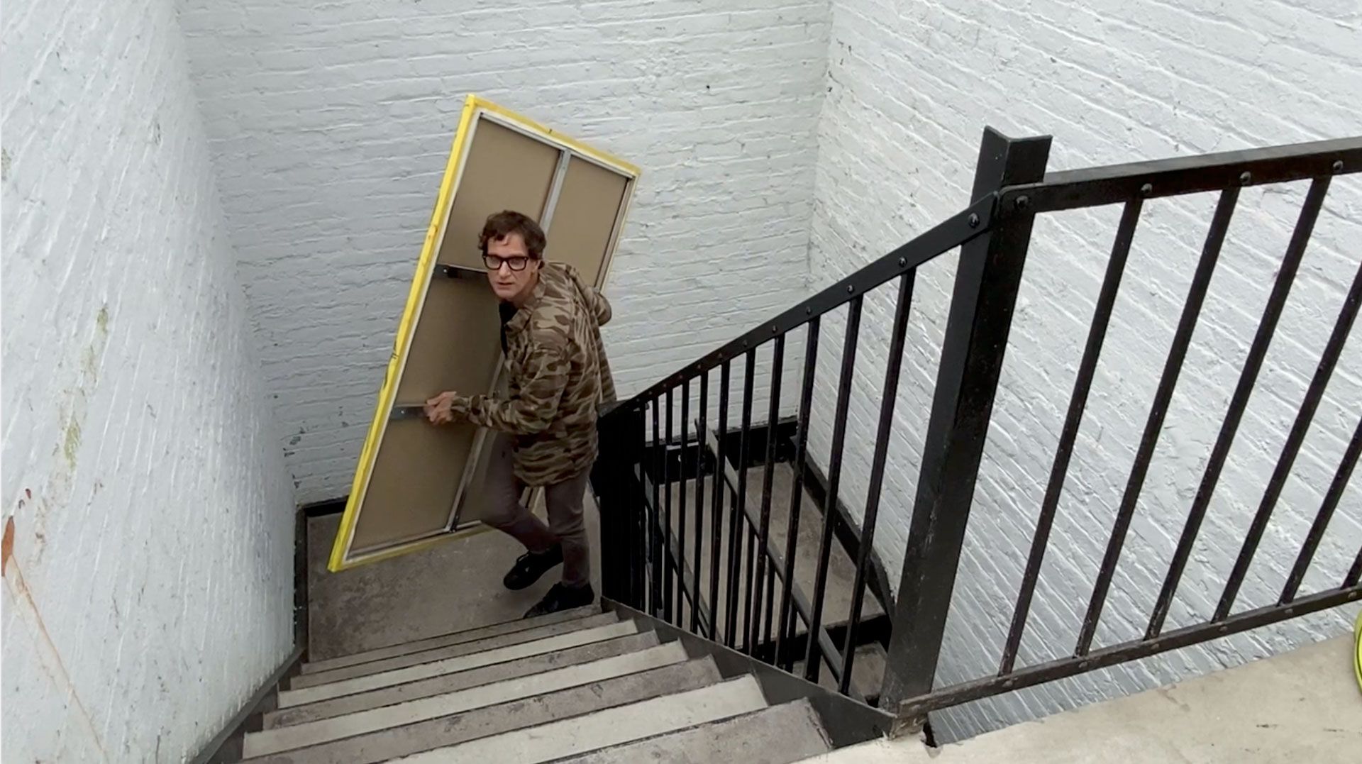A photo of Josh Smith carrying a canvas up the stairs in his studio building, dated 2020.