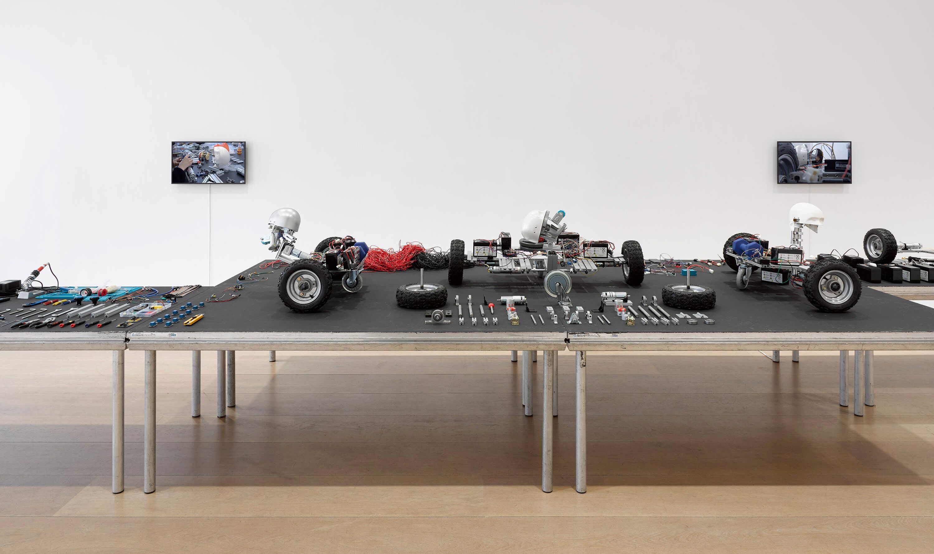 An installation with DIY robotic sculptures made of various materials including aluminum profiles and DC motors, tools and parts arranged on the platform, and the five channel video screens by Geumhyung Jeong, titled Under Maintenance, dated 2021. 