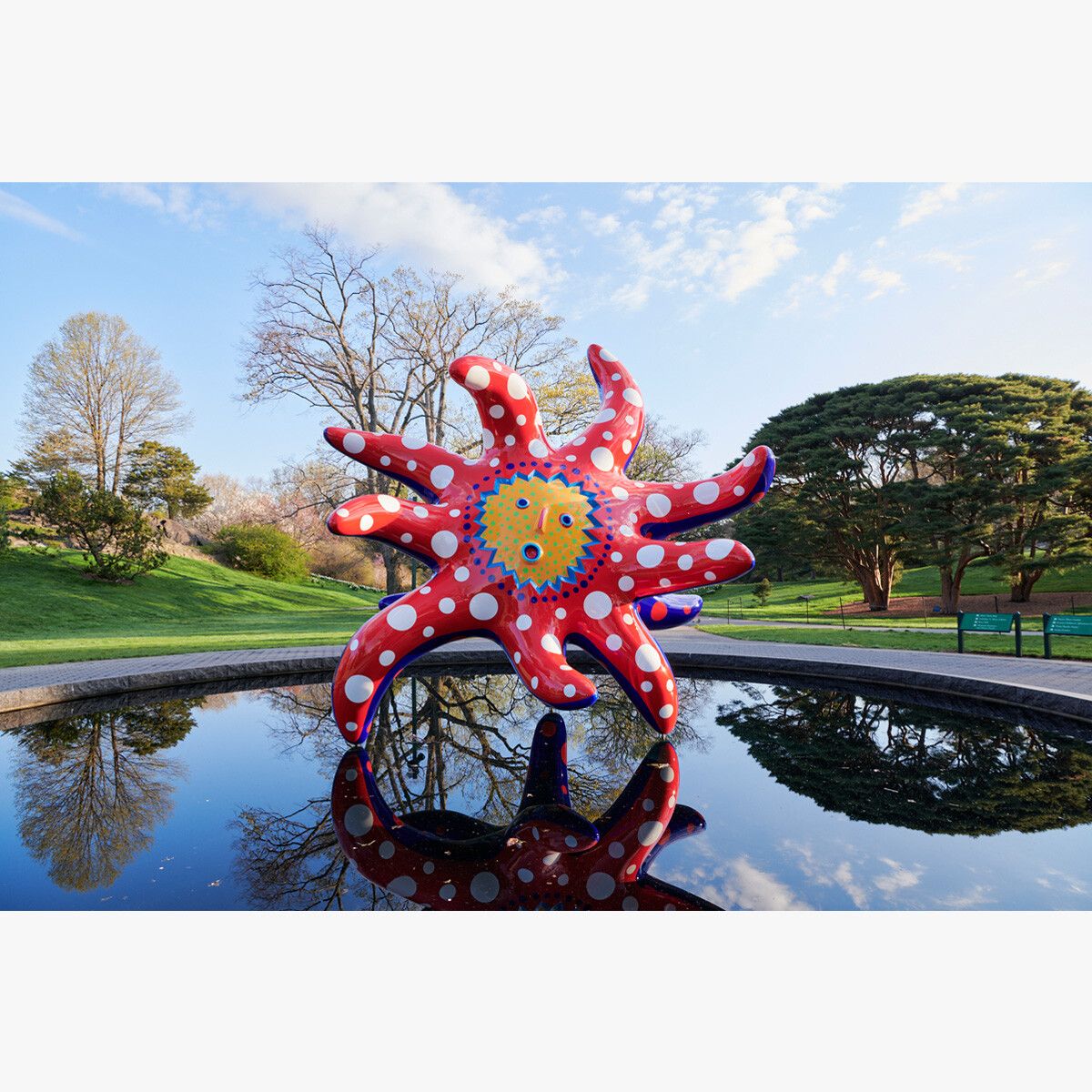 A large tentacled red and blue statue with a yellow sun-like face and white polka dots on its tentacles seems to hover above a reflecting pool, its image mirrored in the water. A mix of trees can be seen in the background.