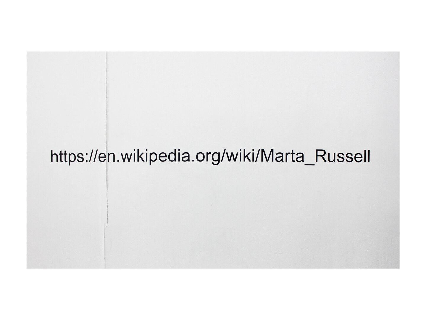 Black vinyl letters on a white wall that read, “https://en.wikipedia.org/wiki/Marta_Russell.” Between the “e” and the “n”, the text crosses over a small crack on the wall.