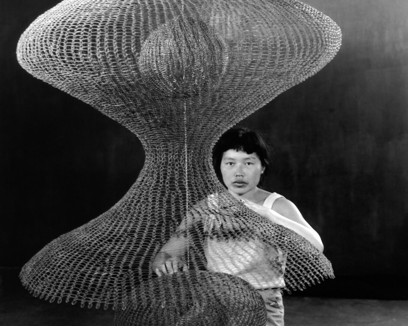 A photograph by Imogen Cunningham of Ruth Asawa kneeling behind a hanging looped-wire sculpture, dated 1957. 