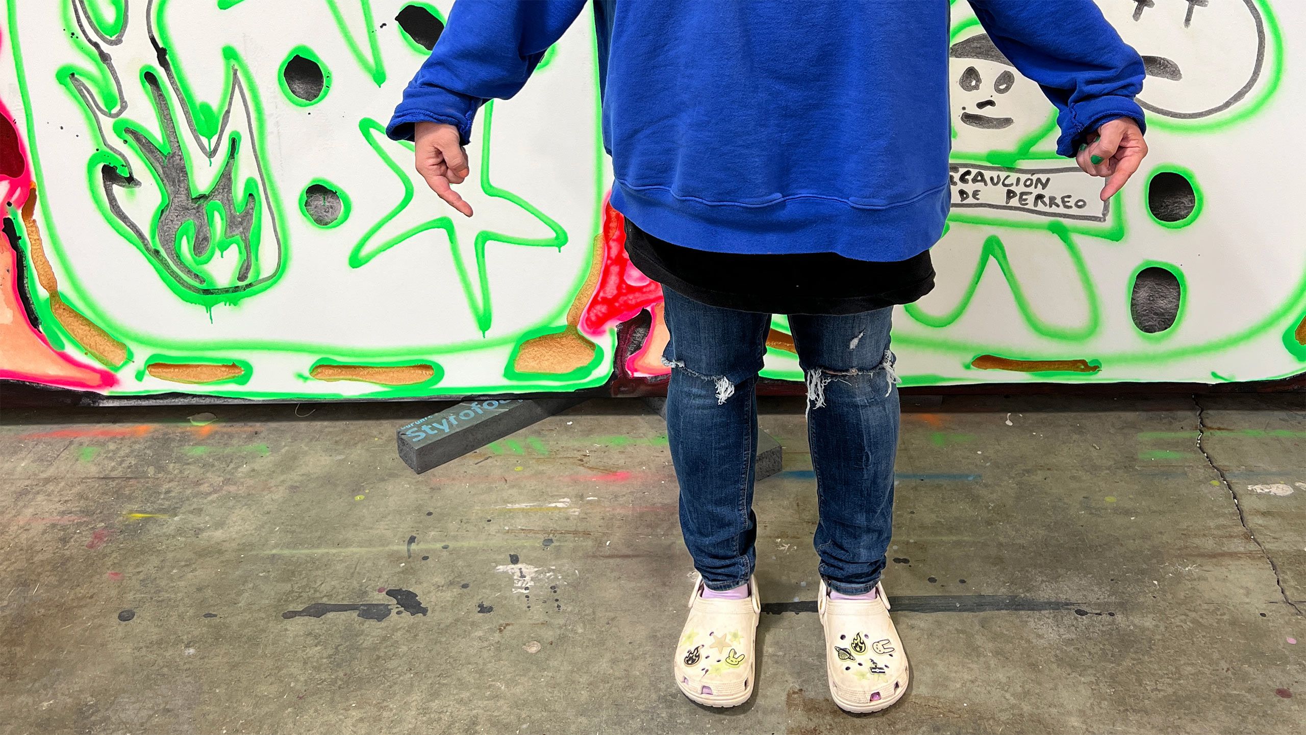 A Photo by Elizabeth Bernhardt  of Katherine Bernhardt in her Bad Bunny x Crocs shoes in front of Crocs World, a large painting that draws from the shoes’ iconography. 