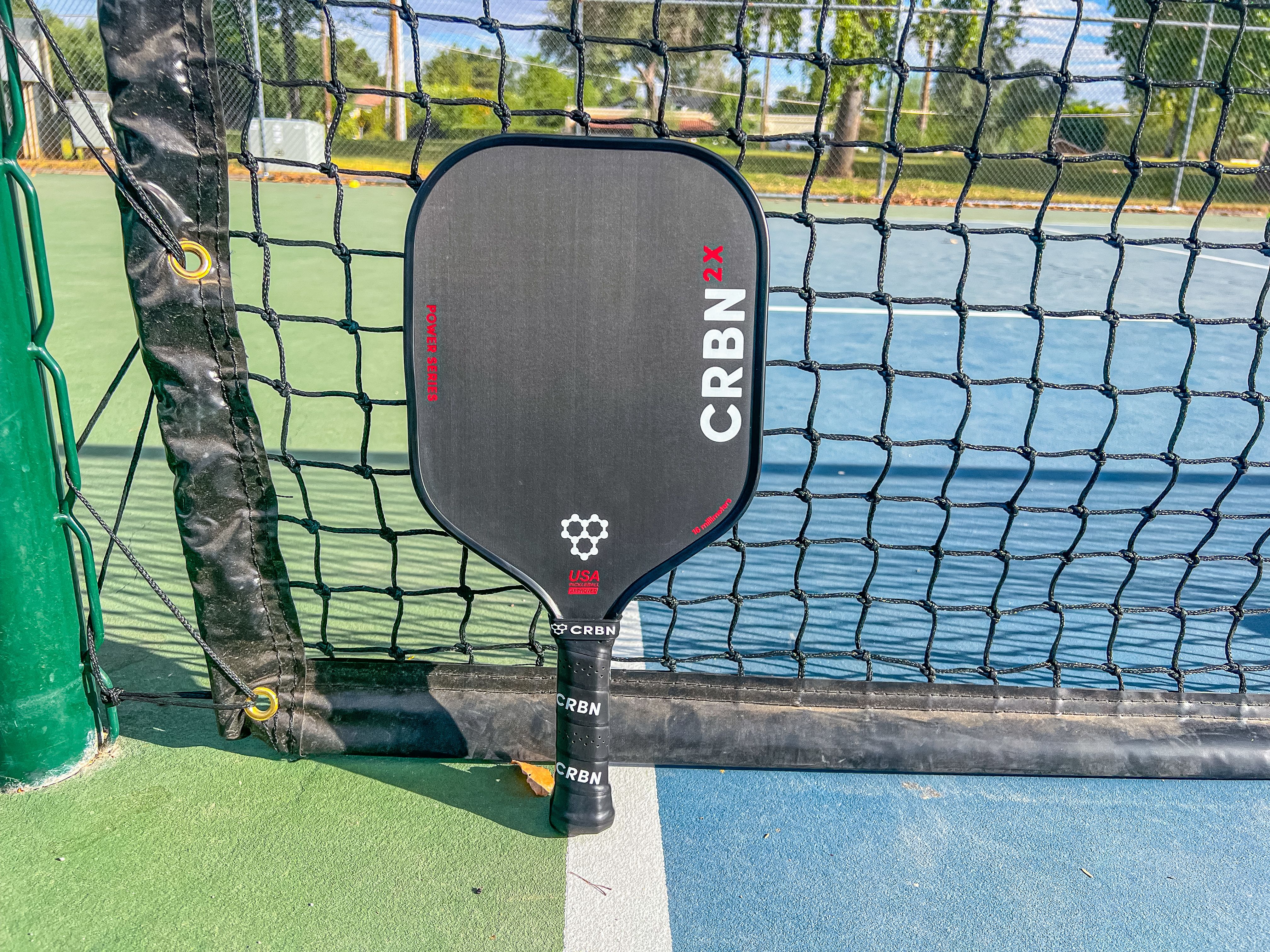 The CRBN-2X Power Series pickleball paddle resting against a net