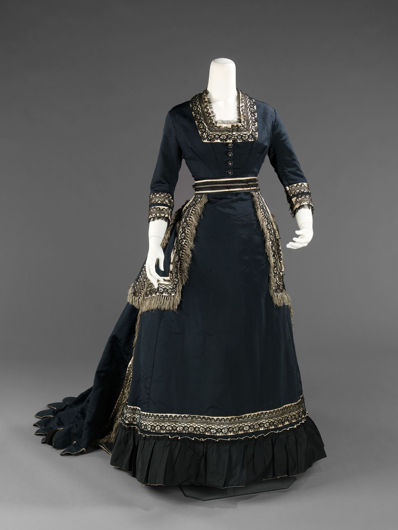 A woman's dress of about 1872-74 in dark blue-black silk on a mannequin, with a square neckline, black lace bands over cream edging at collar, cuffs and hem, fringe down the overskirt, a ruffled hem and a long scalloped train.