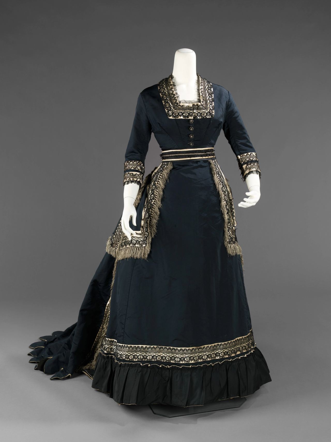 A woman's dress of about 1872-74 in dark blue-black silk on a mannequin, with a square neckline, black lace bands over cream edging at collar, cuffs and hem, fringe down the overskirt, a ruffled hem and a long scalloped train.